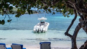 Un bote amarrado en una playa de aguas cristalinas, rodeado de sillas de descanso y un árbol.