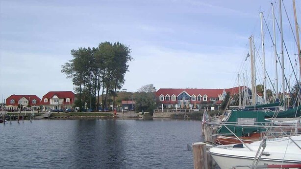 Un puerto deportivo con barcos amarrados, una hilera de casas con techos rojos y una masa de agua tranquila.