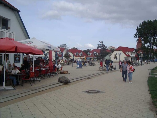 Un espace café en plein air avec des tables et des chaises, quelques personnes qui se promènent et, au fond, un bâtiment avec un toit rouge.