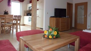 A modern living room with a wooden dining table, red sofas, a TV on a wooden cabinet, and a kitchen in the background. - Ferienhaus Nemo Wohnung 6 "Dorie" (Rerik)