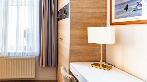 Ein Hotelzimmer mit einer Wand im Holzoptik, einem weißen Schreibtisch mit einer goldenen Lampe, einem gerahmten Bild und einem Vorhangfenster.