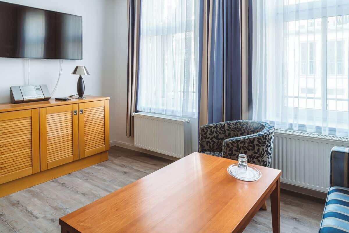 A hotel room with a wooden cabinet, a flat-screen TV, a table with a glass, and a patterned armchair.