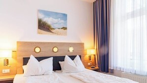 A hotel room with a bed, two bedside lamps, a nightstand, a painting of a beach, and a window with curtains. - Passat (Rostock)