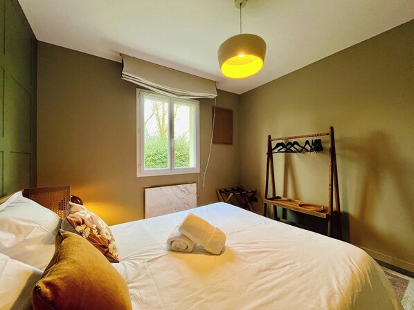 A hotel room with a bed, a window with curtains, a hanging light fixture, and a wooden rack with clothes.