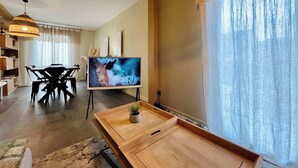 Un soggiorno moderno con zona pranzo, una TV su un mobile e un'ampia finestra con tende leggere.