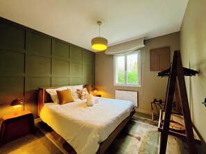 A neatly arranged bedroom with a large bed, a bedside table with a lamp, a window with a view of greenery, and a wooden ladder.
