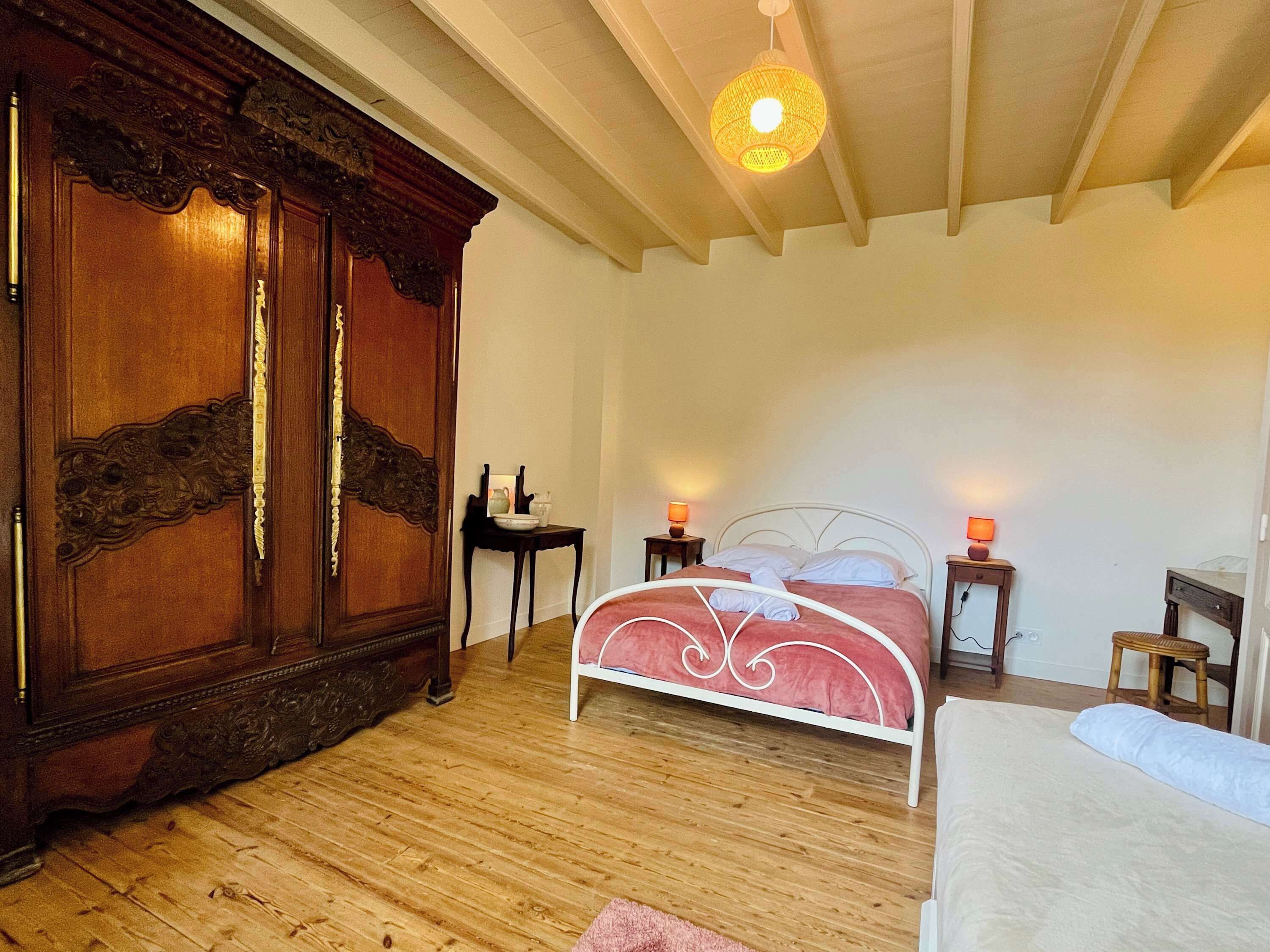 A bedroom with a wooden wardrobe, a bed with white and pink bedding, two bedside tables with lamps, and a hanging light fixture.