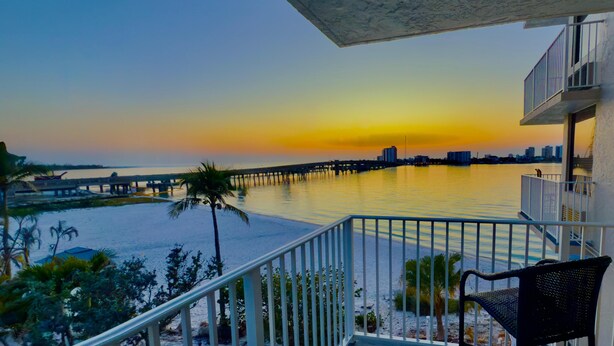 A balcony with a view of a sunset over a body of water, a pier, and a city skyline in the distance. - Charming 1-bedroom apartment with WiFi, AC in fabulous Fort Myers Beach. (Fort Myers Beach)