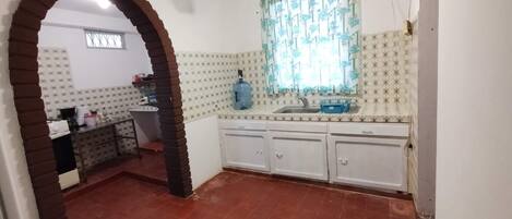 A kitchen with a tiled floor, a window with floral curtains, a ceiling fan, and a water dispenser.
