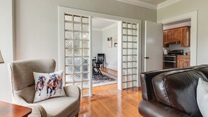 Ein Wohnzimmer mit einem braunen Ledersofa, einem beige farbenen Sessel und einem Holztisch. Eine Glastür führt in einen Essbereich mit einem schwarzen Esstisch und Stühlen. Durch die Glastür ist eine Küche zu sehen.