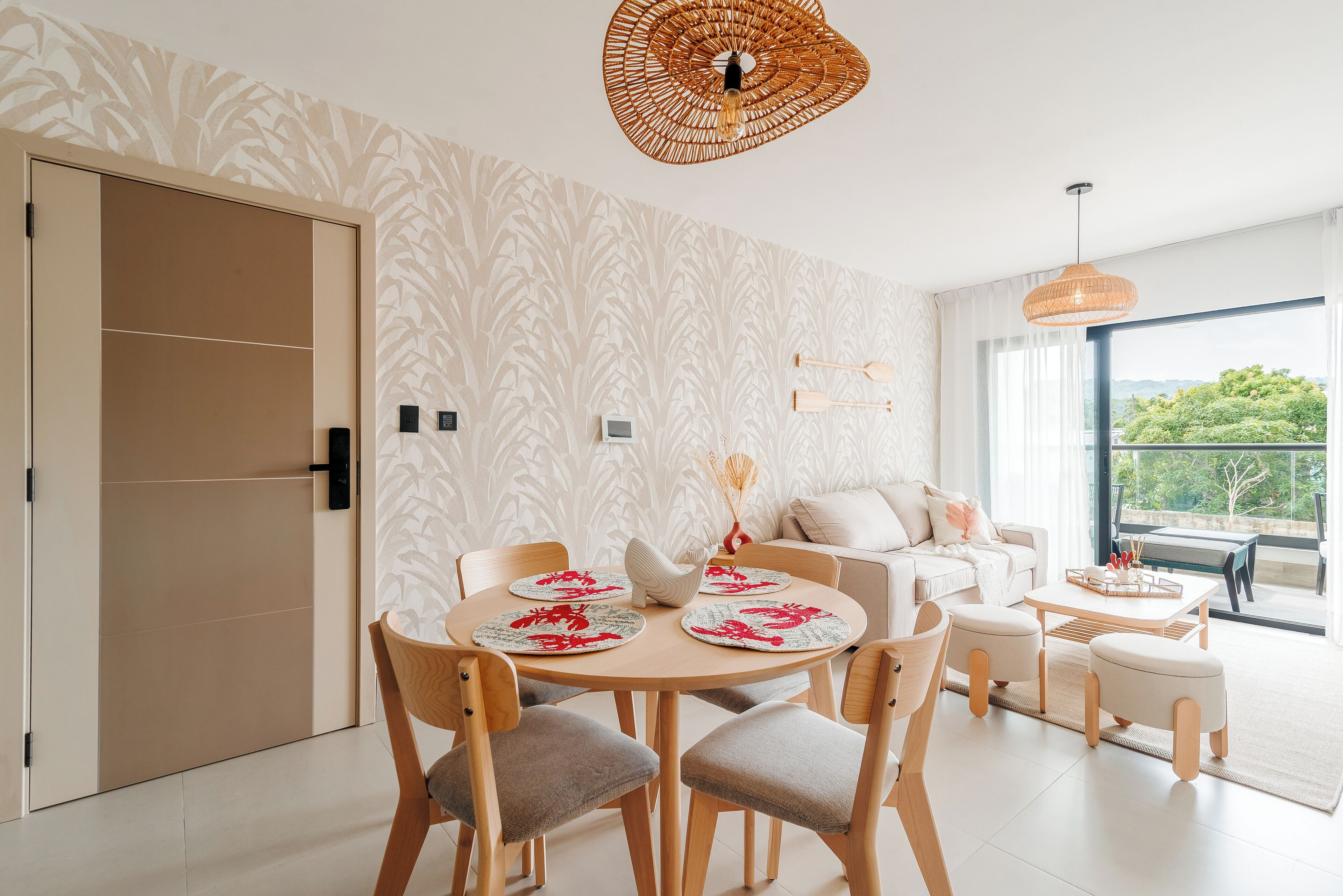 A modern living room with a round dining table, wooden chairs, a sofa, and a large window offering a view of greenery.
