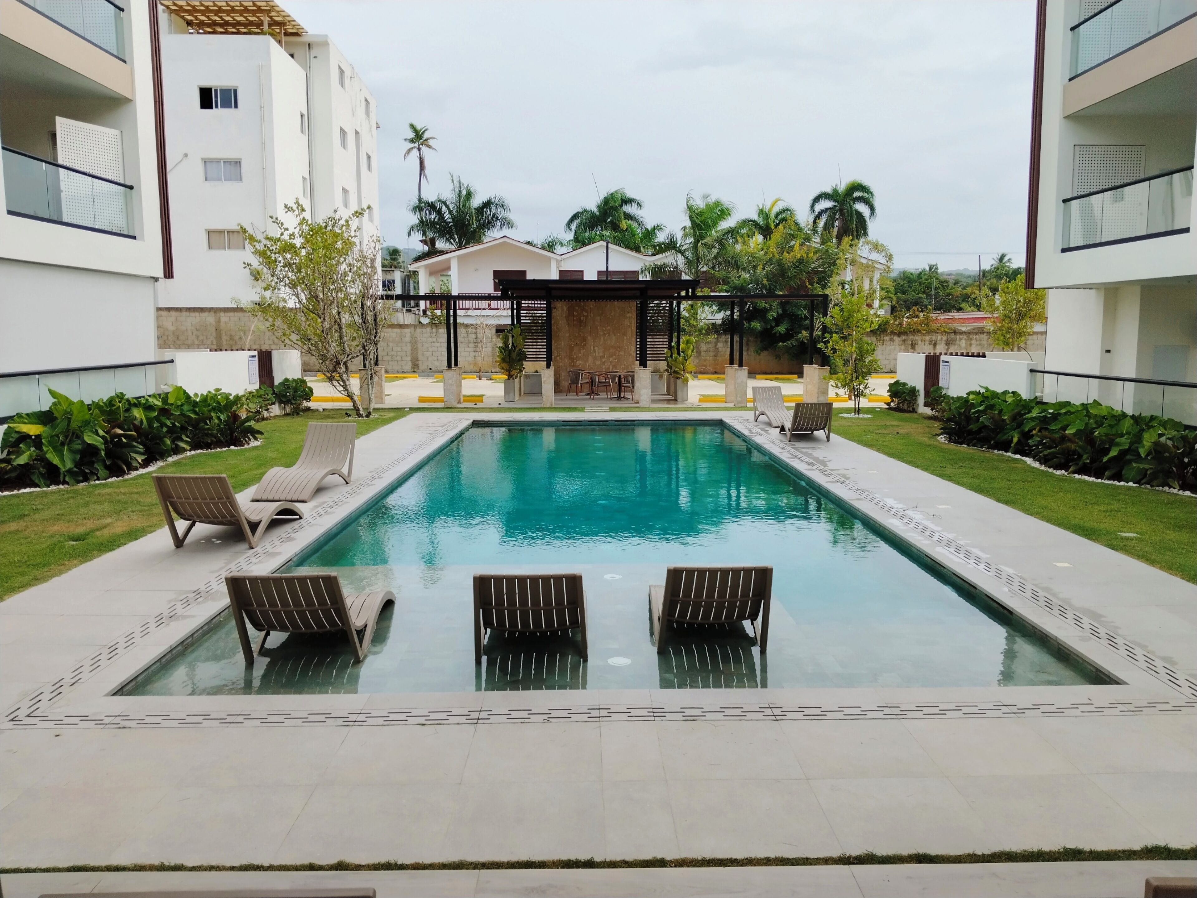 A rectangular swimming pool with lounge chairs and a pergola in a courtyard surrounded by buildings.