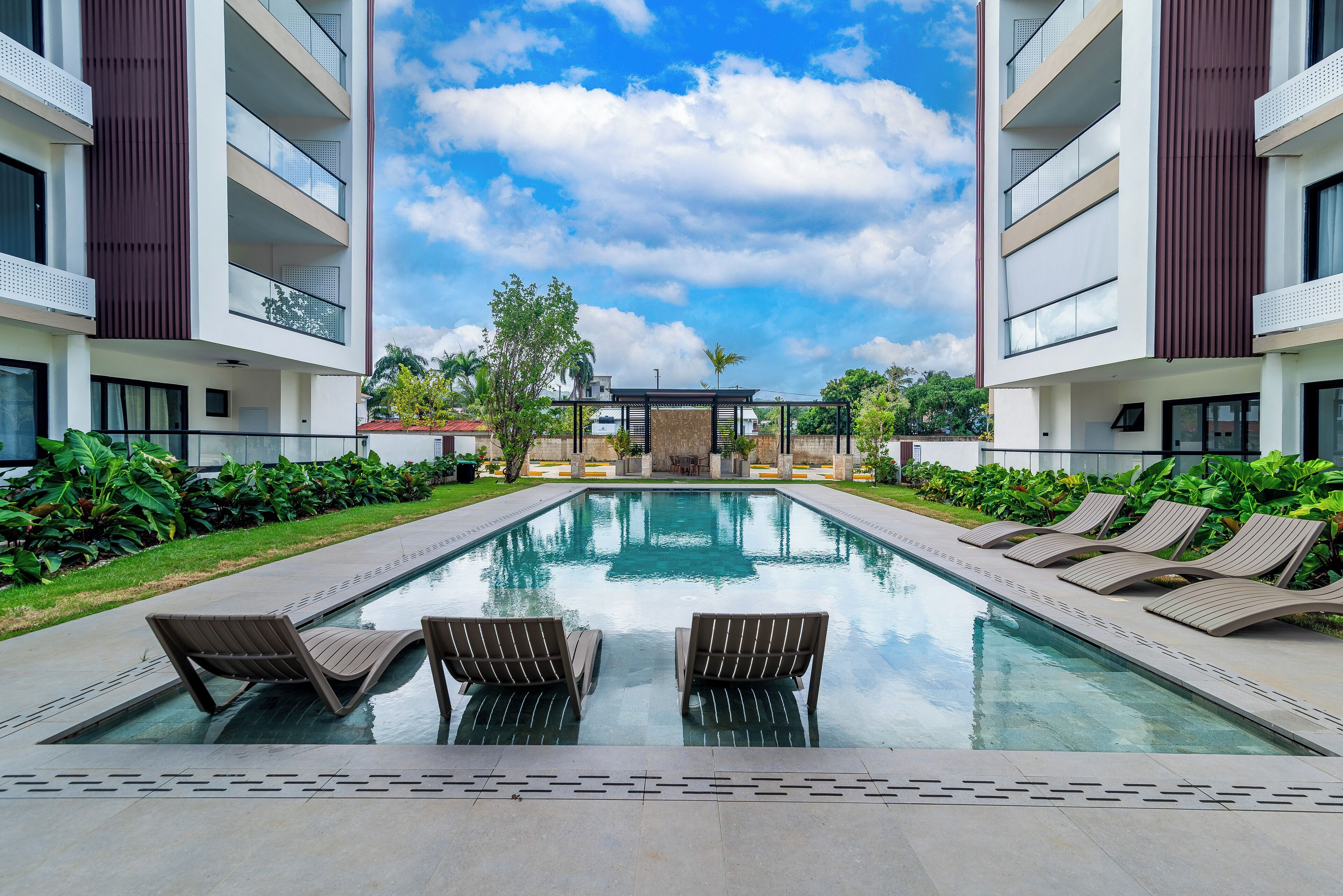 A modern hotel pool area with two lounge chairs facing the pool, surrounded by lush greenery and a clear blue sky.