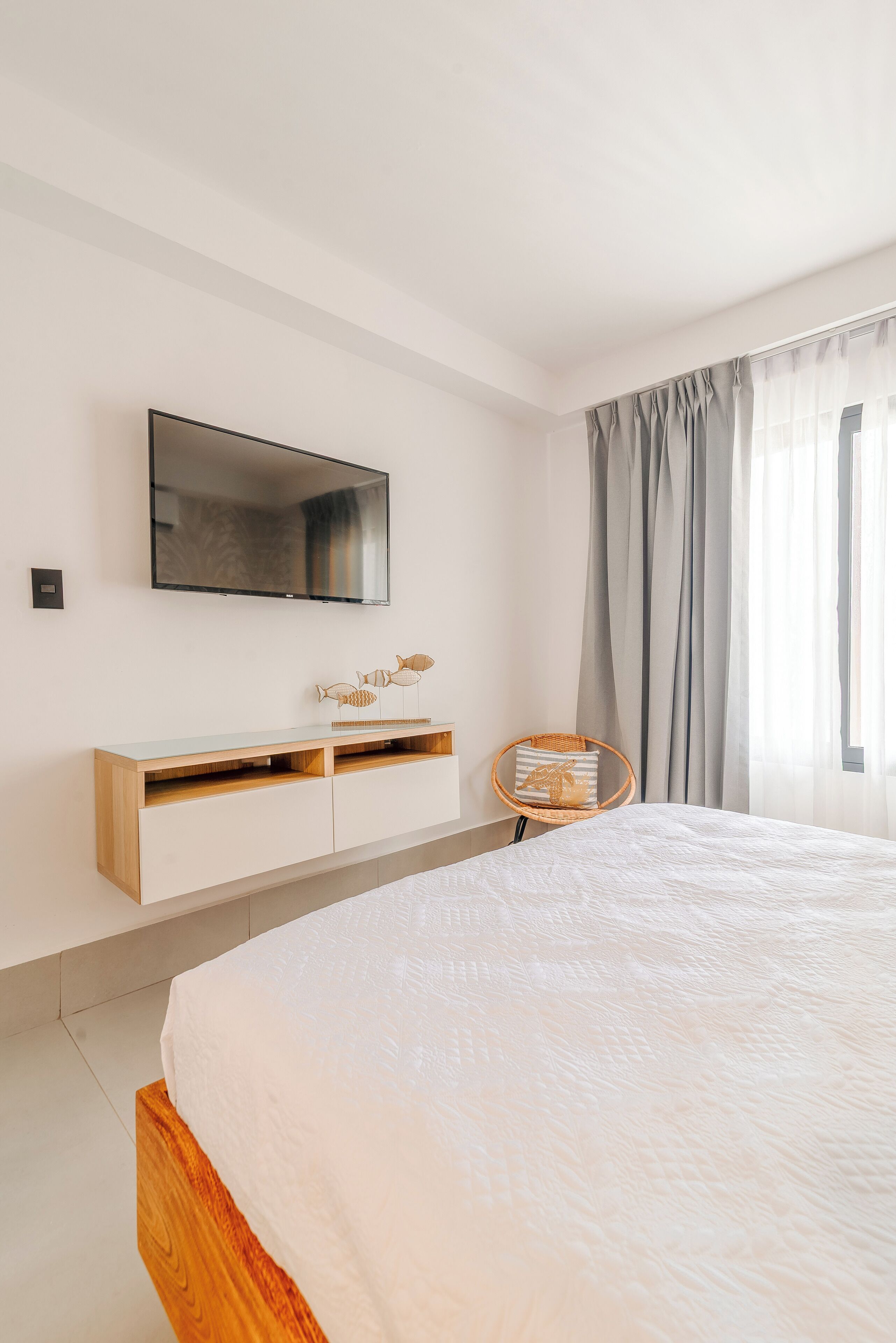 A modern bedroom with a large bed, a mounted TV, a wooden console, and a wicker chair.