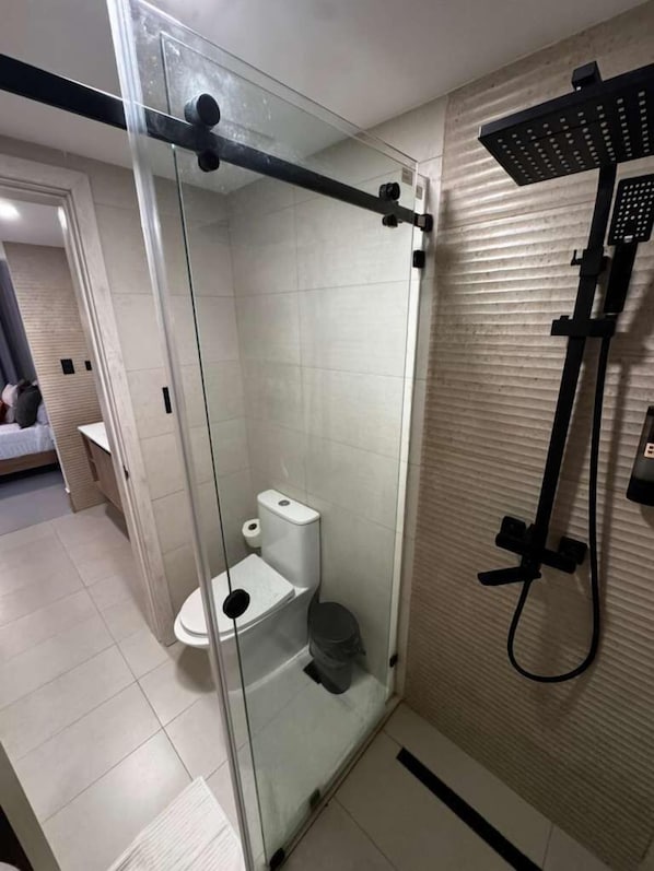 Cabina de ducha con paredes de vidrio, un inodoro, un cabezal de ducha de mano y un perchero para toallas.