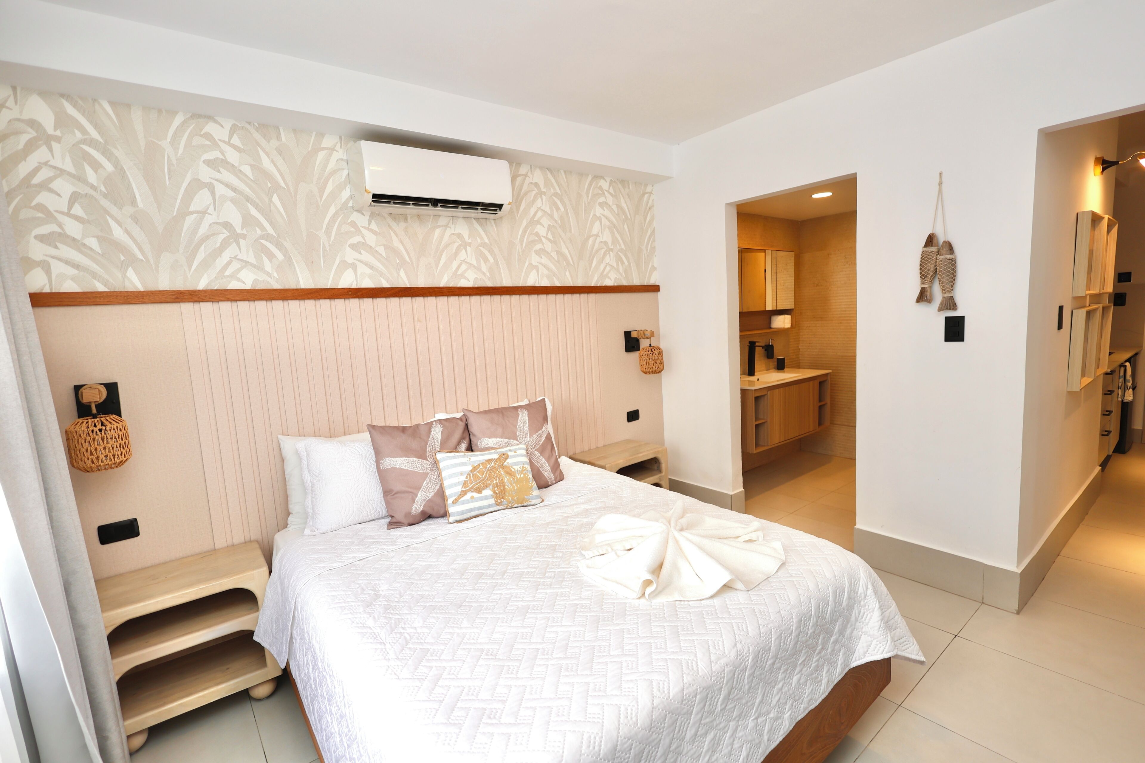 Habitación de hotel ordenada con cama, mesitas de noche, unidad de aire acondicionado, espejo y baño visible a través de una puerta abierta.