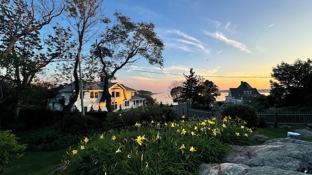 Una casa de techo inclinado, rodeada de un jardín con flores amarillas y vista al mar al atardecer.