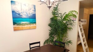 A dining area with a table and chairs, a large painting of a beach scene, a tall green plant, and a white ladder.