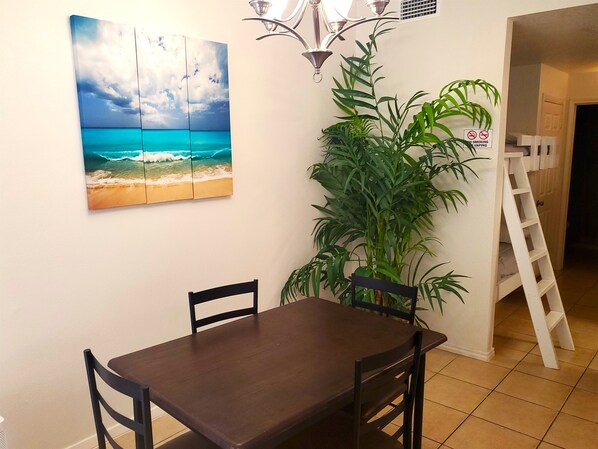 A dining area with a table and chairs, a large painting of a beach scene, a tall green plant, and a white ladder.