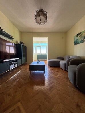 A spacious living room with a wooden herringbone floor, a black coffee table, a grey sofa, and a television on a cabinet.