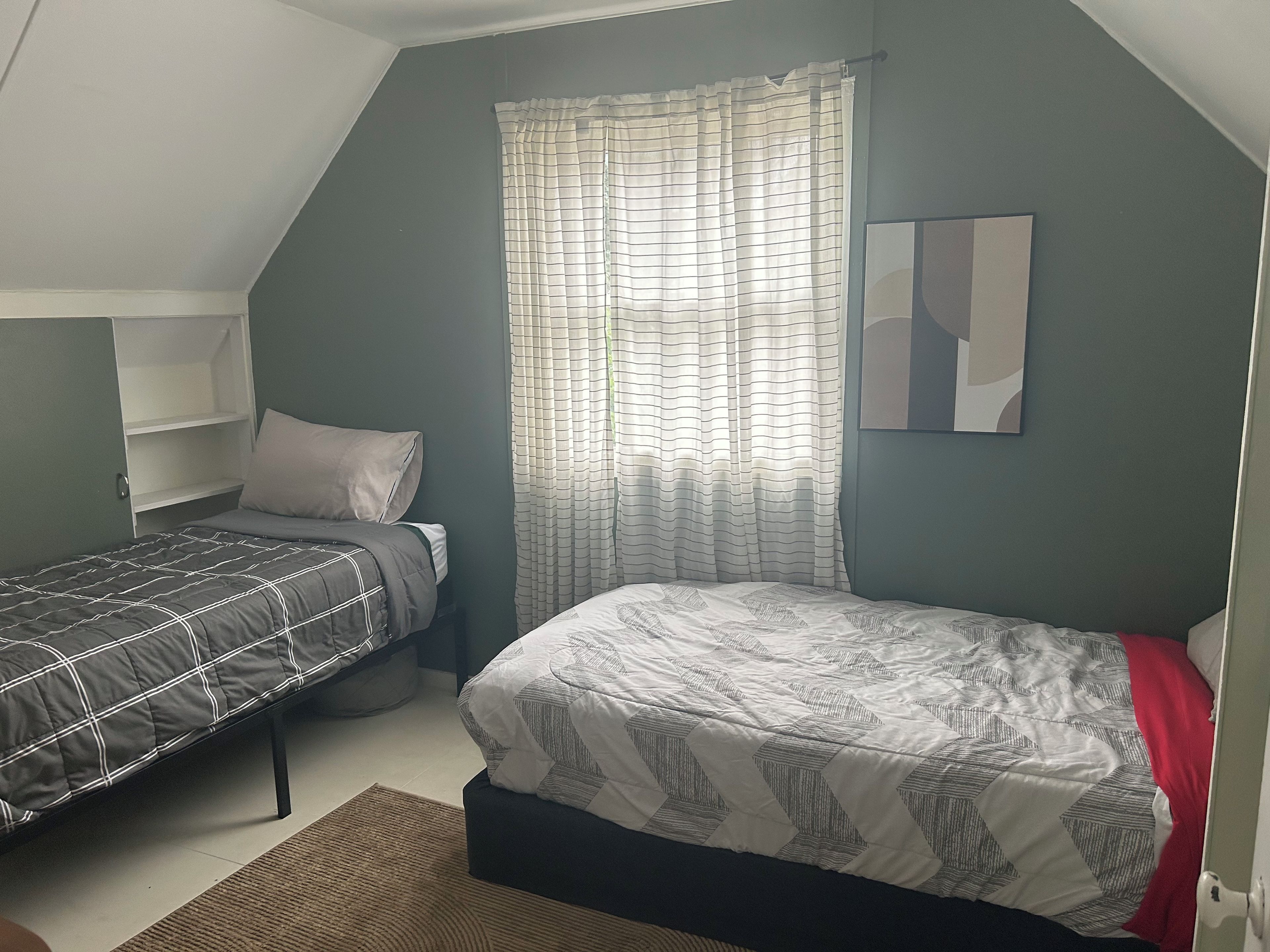 A bedroom with two beds, a window with curtains, a wall-mounted shelf, and a painting.