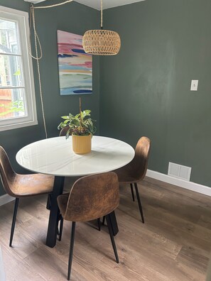 A dining area with a round table, four chairs, a hanging light fixture, and a potted plant.