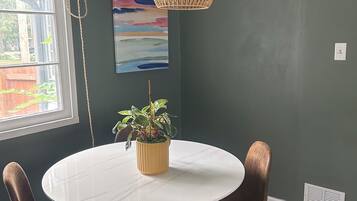 Un comedor con una mesa redonda, cuatro sillas, una lámpara colgante, una planta en maceta y un cuadro en la pared.