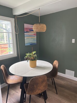 Un comedor con una mesa redonda, cuatro sillas, una lámpara colgante, una planta en maceta y un cuadro en la pared.