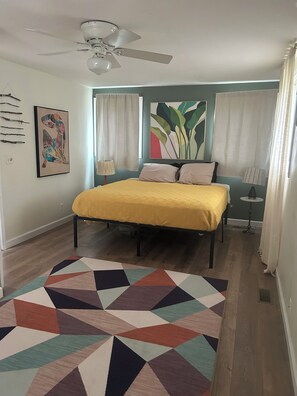 Une chambre à coucher avec un lit, un ventilateur de plafond, un tapis à motifs géométriques et un grand tableau abstrait accroché au mur.