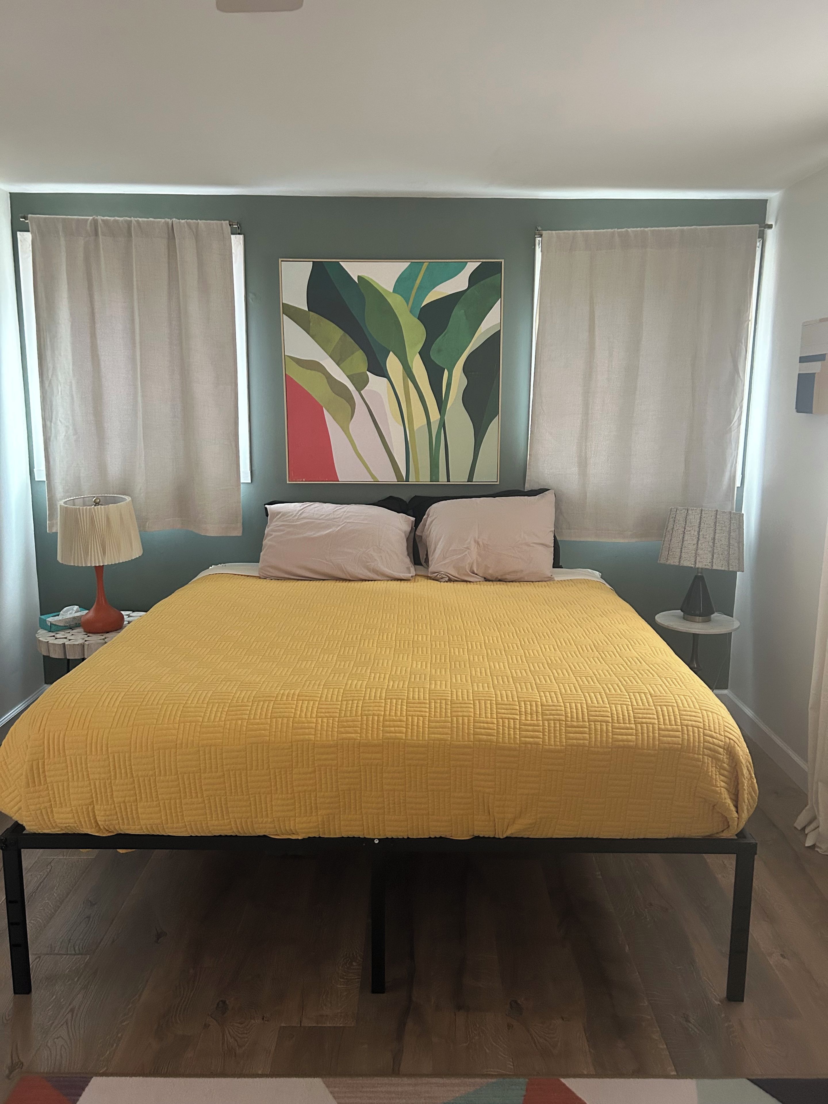 Una cama con colcha amarilla, dos almohadas, una mesita de noche con lámpara y una obra de arte en la pared.