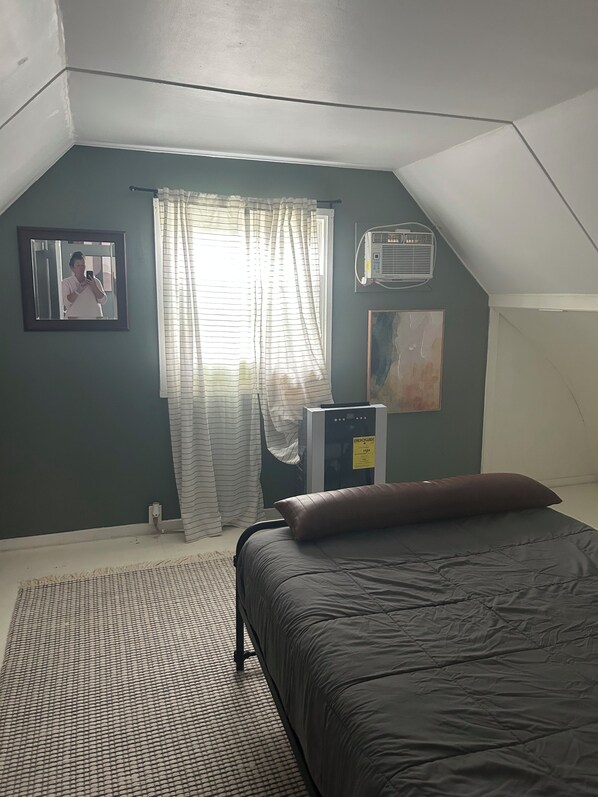 A bedroom with a bed, a window with curtains, an air conditioning unit, and a painting on the wall.