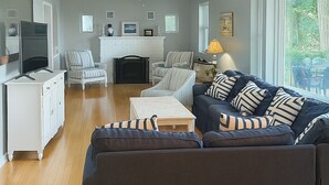 Un salón amplio con un sofá de color oscuro, una chimenea blanca y un ventanal con vistas al exterior.