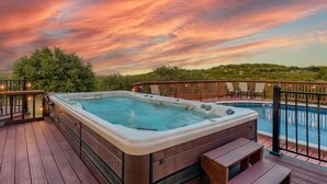 A hot tub on a wooden deck with a pool and chairs, surrounded by a wooden railing and a sunset view.