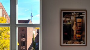 A room with three windows, each offering a different view: one with a building and a tower, another with a building and a tree, and the third with a framed picture of a lamp and a table.