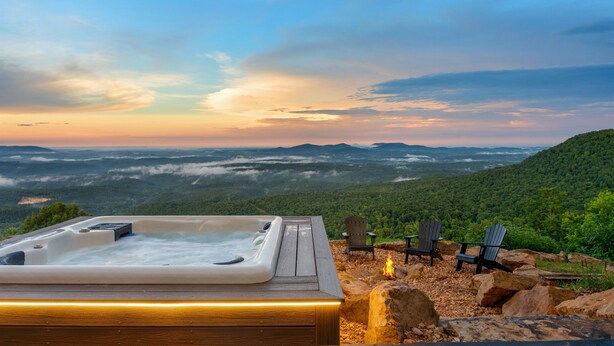 Un jacuzzi en una terraza de madera con sillas y un pozo de fuego, con vistas a un paisaje montañoso.