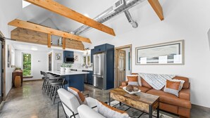 Una cocina moderna con una isla central, un comedor con taburetes de bar, una sala de estar con sofá y mesa de centro, y vigas de madera a la vista.