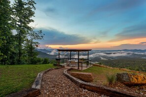 Un pabellón con mobiliario exterior, rodeado por un camino de grava y follaje, con vistas a un paisaje montañoso.