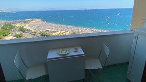 Un balcon avec une table et des chaises, donnant sur une zone côtière où l’on aperçoit des bateaux et, au loin, le paysage urbain.