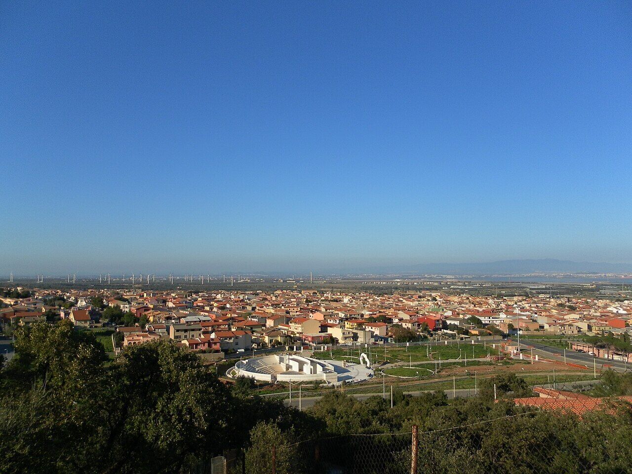 A panoramic view of a town with a mix of residential and commercial buildings, a large open square, and wind turbines in the distance.