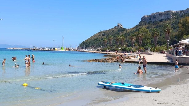 A beach with clear water, people swimming and sunbathing, a lifeguard stand, and a rocky hill in the background.