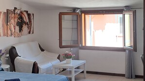 A bedroom with a bed, a bedside table with a vase of flowers, a window with a view, and a wall with artwork.