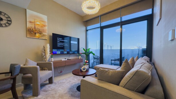 A modern living room with a large TV, comfortable seating, a coffee table, and a view of the city through large windows.