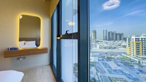 A modern hotel room with a large window, a bedside table with a lamp, and a view of the cityscape.