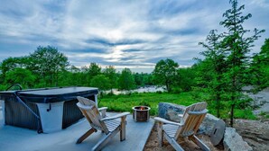 An outdoor seating area with two Adirondack chairs, a hot tub, and a fire pit, overlooking a natural landscape.