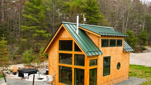 A small wooden cabin with a green roof, large windows, and a gravel driveway.