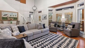 Un ampio soggiorno con un grande divano grigio, due poltrone e un tappeto decorato. Diverse finestre offrono vista su alberi e il soffitto è in legno.