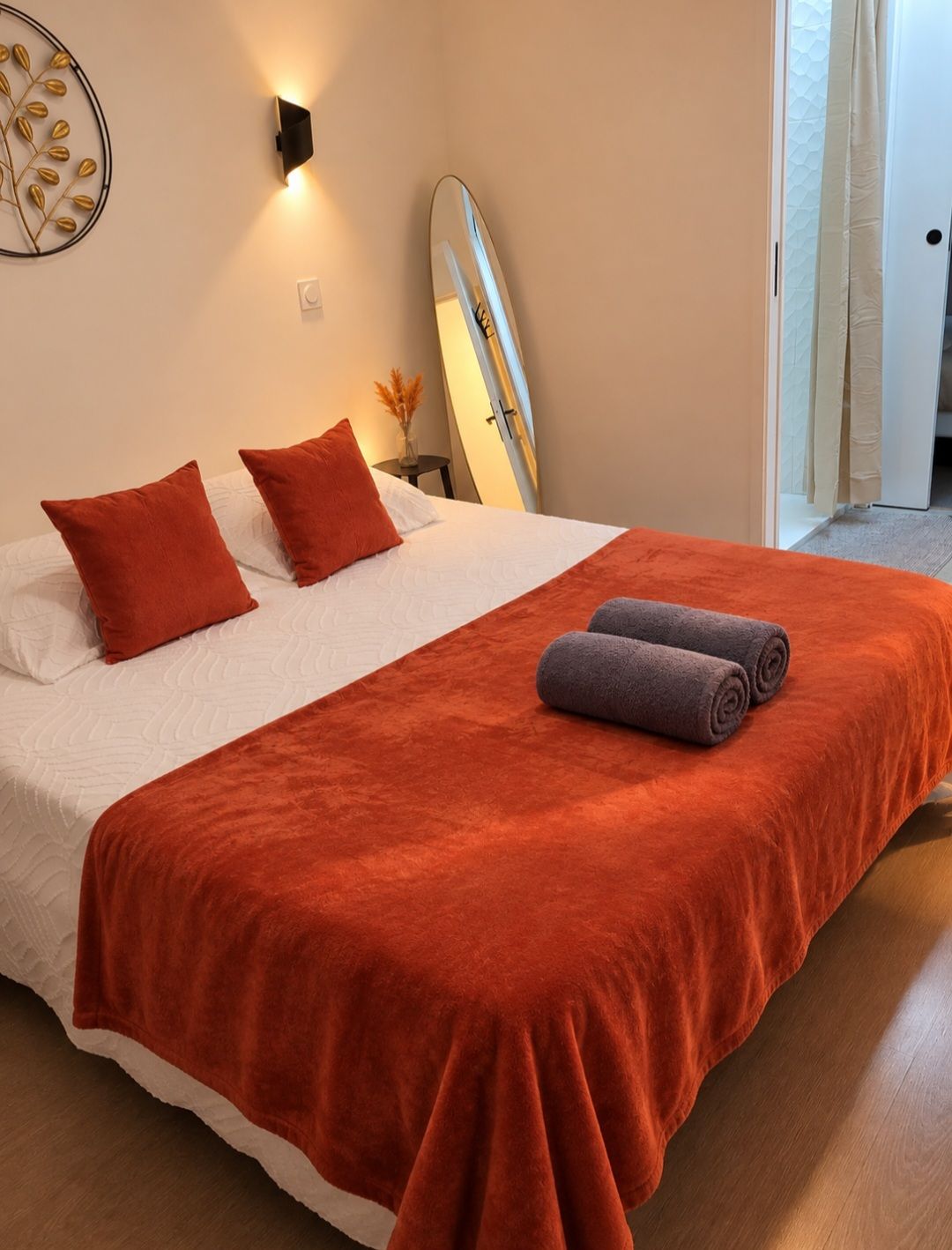 Een net opgemaakt bed met een rode deken en kussens, een wandlamp en een surfboard dat tegen de muur staat.