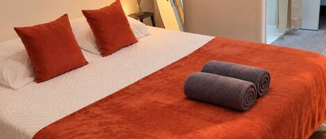 A neatly made bed with a red blanket and pillows, a wall-mounted light, and a surfboard leaning against the wall.