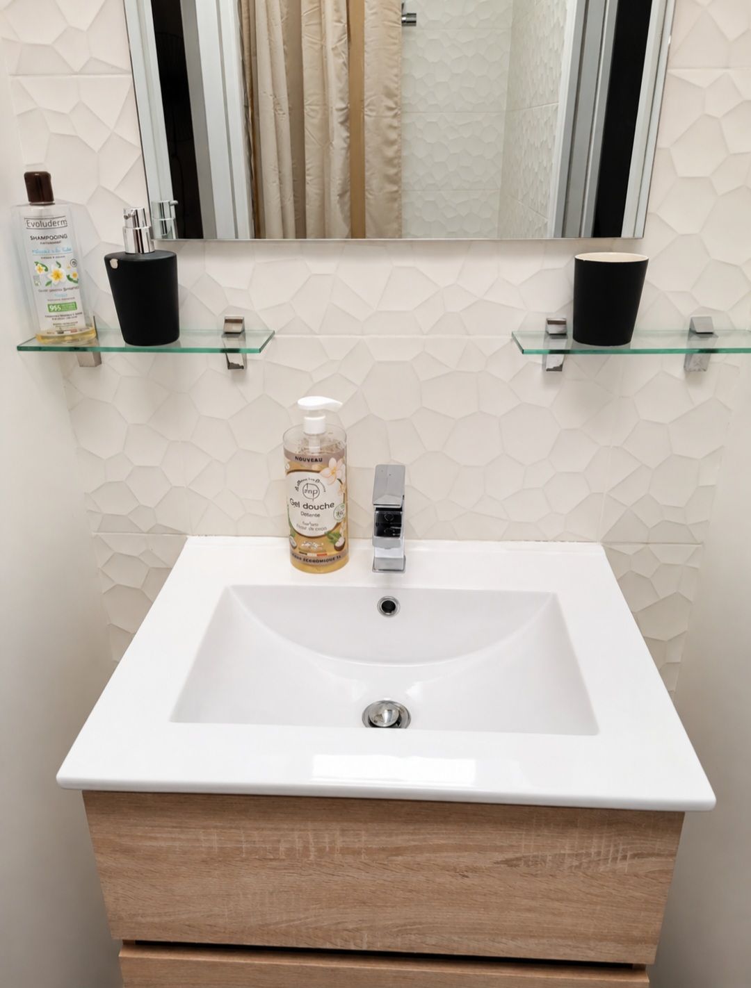 Une salle de bain avec un lavabo blanc, un miroir au-dessus, et deux étagères en verre où sont rangés les articles de toilette.