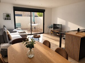 A modern living room with a wooden dining table, a sofa, a TV, and a sliding glass door leading to an outdoor patio.